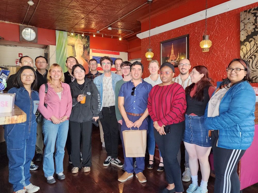 16 members of the DAISY research lab stand in small arc inside a Thai restaurant smiling and facing the camera. In the center of the photo are the people of honor Dr. Ricardo Flores holding a gift bag and Dr. Walter Gerych.